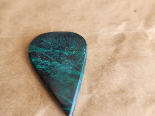 A hand-polished 54-carat Eilat stone cabochon with deep green and blue swirling patterns on a brown paper background. קבודון אבן אילת במשקל 54 קארט, מלוטש ידנית משני הצדדים, בעל דוגמאות בגווני ירוק וכחול על רקע נייר חום.