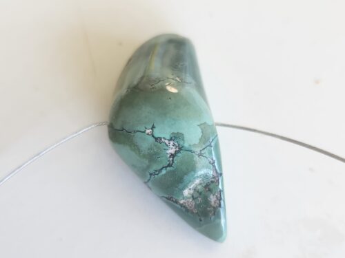 A smooth, 60-carat Tibetan turquoise bead with dark matrix veins, strung on a thin wire. חרוז טורקיז טיבטי 60 קראט במרקם חלק עם ורידים כהים, מושחל על חוט דק.